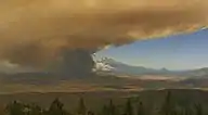 Thick orange-brown smoke blocks half a blue sky, with conifers in the foreground