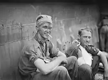 A man with a bandage around his head, smoking a cigarette beside another man with his right arm in a sling