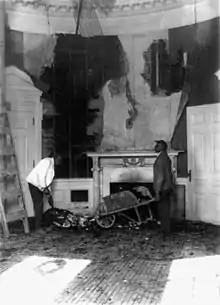 a black and white image of a room ravaged by fire with a man holding a wheelbarrow full of debris in front of a fireplace