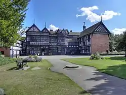 Photo a long, two-storeyed house with exposed wooden beams, many gables and small-paned windows. In the foreground is a lawn and trees are on the left.