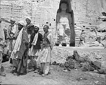 Black and white photograph of 6 Afghan men. Behind them, a giant sculpture is carved into the mountain side.