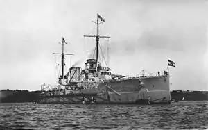 Close front oblique view of a gray warship with two funnels, two visible gun turrets, and two masts