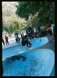 A person does an airborne trick between two light blue bowls in the skate park in Owl's Head. A few people watch from above the bowl.