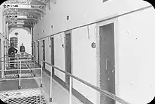 "Black and white photograph of a landing in Kilmainham Jail, with closed cell doors visible on either side and two uniformed guards standing towards the left"