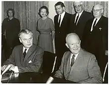 Diefenbaker and a smiling bald man in a suit sit at a table.  Two women and two men stand behind them.