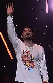 Jon Bellion lifting his right arm, wearing a white long sleeve shirt.