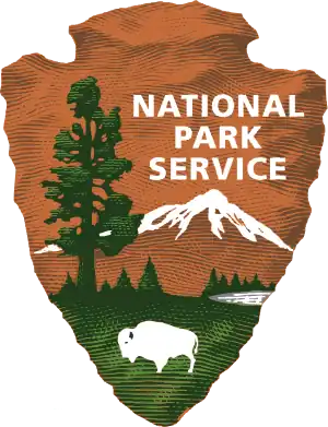 The National Park Service logo depicts an outline of a brown, jagged arrowhead, embedded in which are the silhouettes of a large green tree, a white mountaintop, and a grazing white bison. The words "National Park Service" hover above the mountaintop.