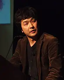 A Japanese man in a brown jacket and dark grey shirt standing at a podium.