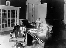 A black and white photo of desk and chair. A thick black ribbon encircles the chairs with a bow and another ribbon does the same around the central part of the desk.