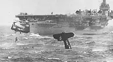 The dark tail of a Navy plane bobs upright in foggy, choppy seas, while a low hovering twin-rotor helicopter pulls a pilot from the water with a rope. The parent aircraft carrier just behind, with a handful of crew seen standing on the edge of the deck watching.
