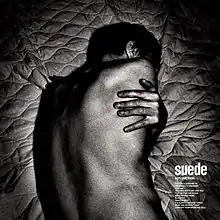 A stark black and white image of a man lying on a mattress. His back is facing the camera and his face is obscured. His hand is resting on his back as if his arms are wrapped around him.