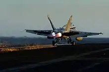 Photograph from behind a twin-engined jet fighter. The aircraft's wheels are on the surface, but the engines are still active, and a hook on the underside of the aircraft is in contact with the surface and trailing sparks