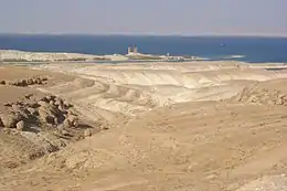 Two weathered stone towers in the background partly surrounded by the water of a river, while mounds of desert sand are pictured in the foreground