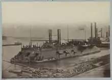 A ship along a shoreline.  The vessel has two smokestacks, flies the American flag, and multiple cannon are visible.